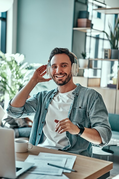 Feliz empresário em fones de ouvido ouvindo música enquanto está sentado no café Homem positivo trabalhando com laptop e documentos dentro de casa