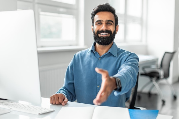 Feliz empresário indiano oferecendo mão para aperto de mão sentado na mesa de escritório com computador sorrindo para