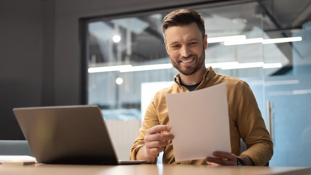 Feliz empresário lendo papéis de negócios sentado perto de laptop no escritório