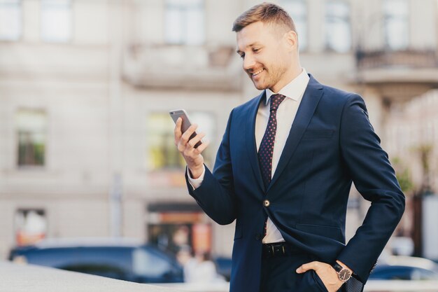 Feliz empresário mantém a mão no bolso, vestindo terno formal e relógio de pulso e usando telefone inteligente