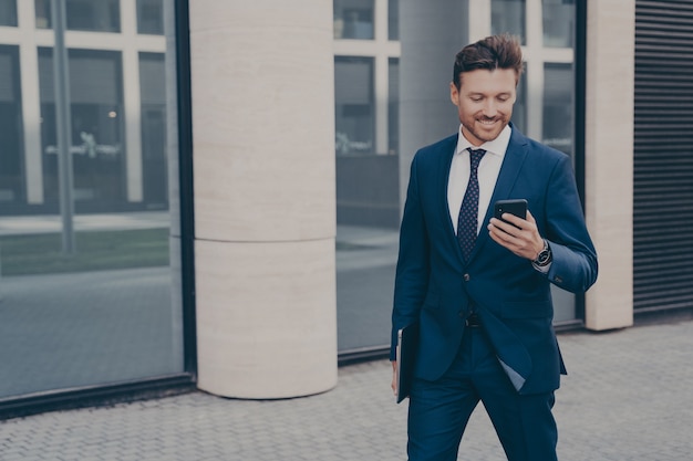 Feliz empresario masculino europeo en traje sosteniendo teléfono móvil y sonriendo al aire libre