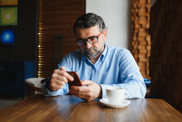 Feliz empresário sentado no restaurante e esperando o almoço Ele está usando telefone inteligente e conversando com alguém Conceito de estilo de vida de idosos de negócios