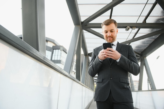 Feliz empresário sorridente vestindo terno preto e usando smartphone moderno perto do escritório no empregador bem-sucedido de manhã cedo para fazer um acordo em pé perto do escritório do arranha-céu à luz do sinalizador noturno