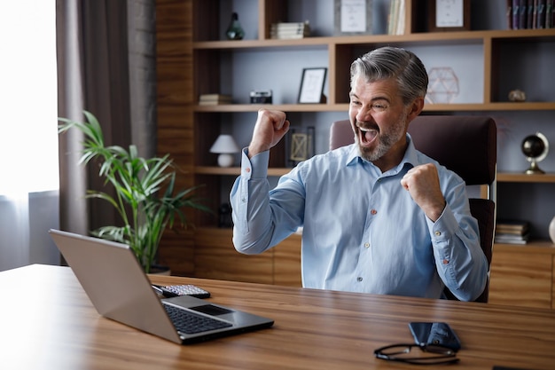 Feliz empresário surpreso com as boas notícias e fazendo um gesto de sim enquanto olha para o laptop sentado no escritório Comerciante desfrutando de sucesso comemorando a vitóriafaça um bom negócio obter lucro