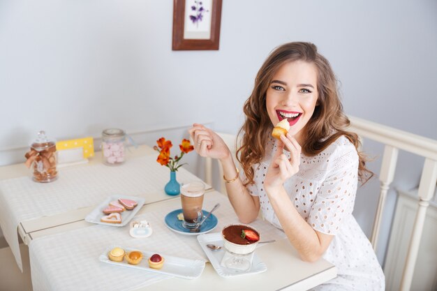 Feliz encantadora joven comiendo pasteles pequeños y bebiendo café con leche en el café