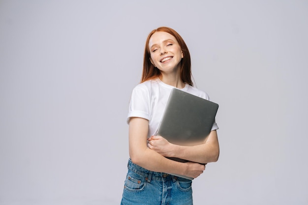 Feliz y encantadora joven estudiante sosteniendo una computadora portátil con los ojos cerrados en un fondo gris aislado. Bonita modelo de dama con el pelo rojo que muestra emocionalmente expresiones faciales en el estudio, copie el espacio.