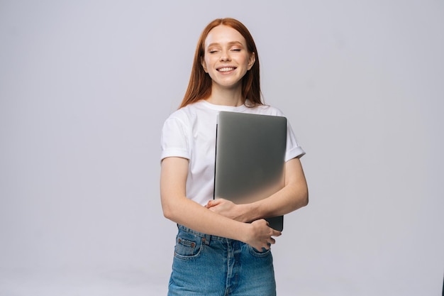 Feliz y encantadora joven estudiante sosteniendo una computadora portátil con los ojos cerrados en un fondo gris aislado. Bonita modelo de dama con el pelo rojo que muestra emocionalmente expresiones faciales en el estudio, copie el espacio.