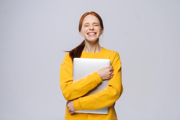 Feliz encantadora joven estudiante sosteniendo una computadora portátil con los ojos cerrados sobre un fondo gris aislado Bonita dama modelo con el pelo rojo mostrando emocionalmente expresiones faciales en el espacio de copia del estudio