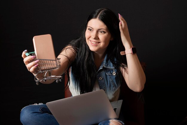 Foto feliz y encantadora mujer joven con carrito de compras y teléfono inteligente usando una computadora portátil para comprar en línea