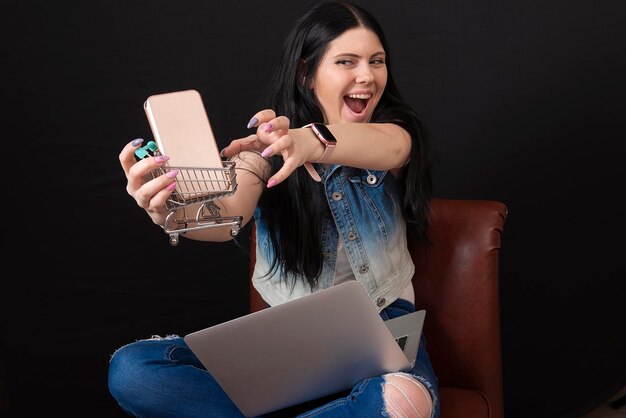 feliz y encantadora mujer joven con carrito de compras y teléfono inteligente usando una computadora portátil para comprar en línea