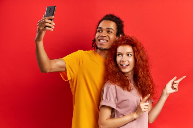 Una feliz y encantadora pareja de raza mixta ha paseado juntos, se paran uno al lado del otro, posan para hacerse selfie, están de buen humor. Diversos amigos toman fotos de sí mismos, pared roja. Los turistas van de turismo.