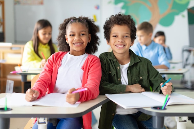 Feliz escolar negro y estudiante mirando a la cámara y sonriendo sentados en los escritorios con otros