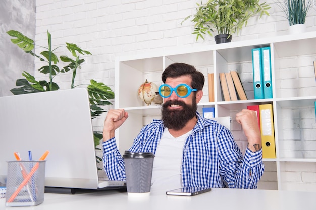 Feliz especialista em computadores em óculos extravagantes faz gesto de vencedor olhando para monitor de laptop no poder de escritório moderno