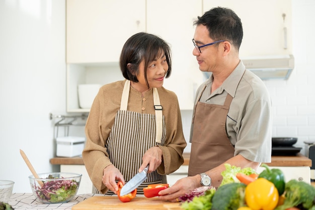 Una feliz esposa asiática está cortando un pimienta dulce mientras su marido la ayuda en la cocina