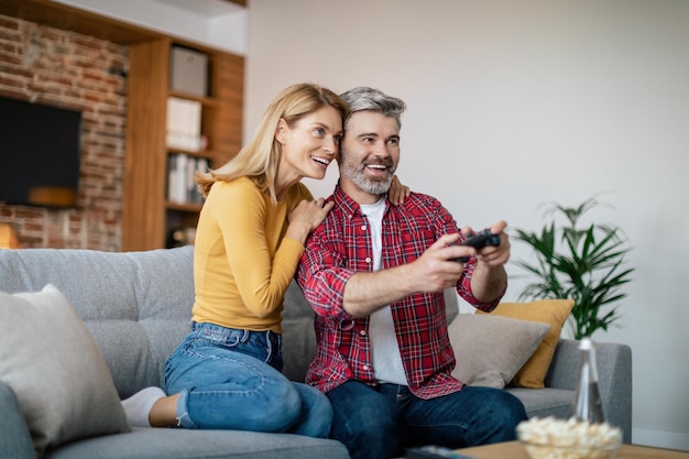 Feliz esposa europea madura abrazando a su marido con joystick disfruta del juego en línea en la computadora sentada en el sofá