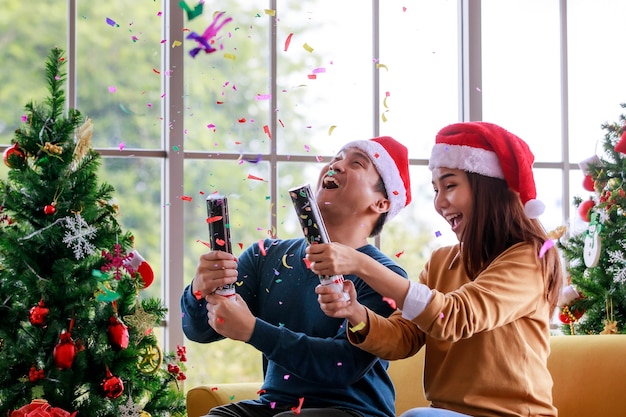 Feliz esposo y esposa de la familia asiática viste un suéter con sombrero rojo y blanco de santa claus sentado en un sofá amarillo tirando de confeti de papel brillante de popper de fiesta explosivo juntos celebrando la víspera de Navidad.