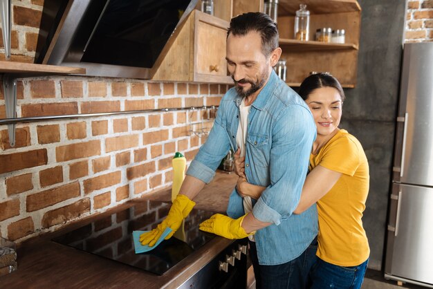 Foto feliz esposo joven barbudo limpiando la cocina mientras su esposa sonriendo y abrazándolo