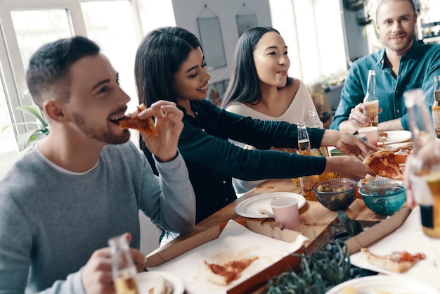 Feliz de estar cerca. Grupo de jóvenes en ropa casual comiendo pizza y sonriendo mientras tienen una cena en el interior