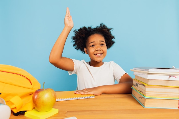 Feliz estudante afro-americana sentada na mesa na aula e esticando a mão para cima no fundo azul de volta ao conceito de escola