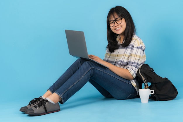 Feliz estudante asiática sentada usando laptop em um fundo azul