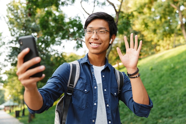 Feliz estudante asiático do sexo masculino em óculos fazendo selfie no smartphone e acenando para a câmera enquanto está no parque