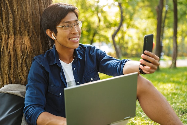 Feliz estudante asiático do sexo masculino em óculos fazendo selfie no smartphone e segurando o laptop enquanto está sentado perto da árvore no parque