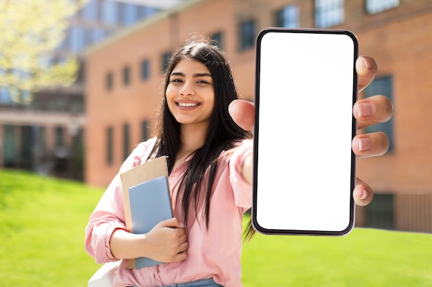 Feliz estudante hispânica mostrando smartphone com tela vazia e sorrindo enquanto posava ao ar livre