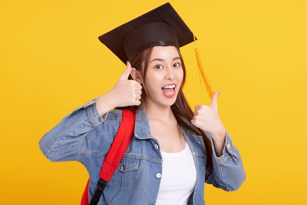Feliz estudante universitária asiática em chapéu de formatura com gesto de polegar para cima