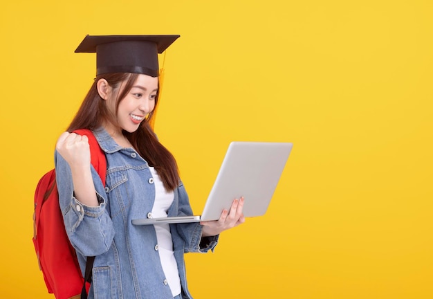 Feliz estudante universitária asiática em chapéu de formatura e segurando laptop