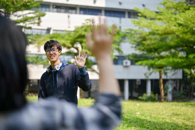 Feliz estudante universitário asiático acena com a mão para cumprimentar seu amigo no parque do campus