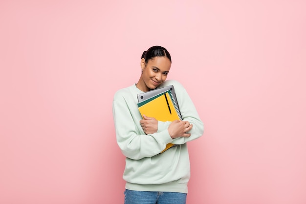 feliz estudiante afroamericano en sudadera sosteniendo portátil y cuadernos aislados en rosa