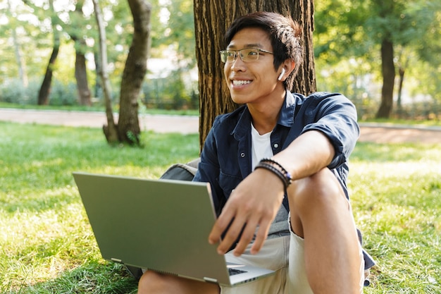 Feliz estudiante asiático en anteojos con computadora portátil mirando a otro lado mientras está sentado cerca del árbol en el parque