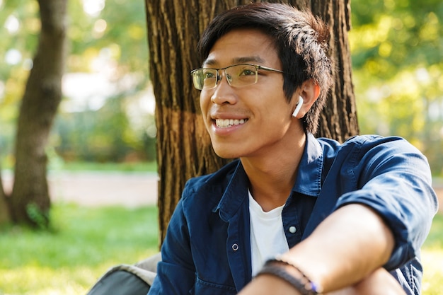 Foto feliz estudiante asiático en anteojos mirando a otro lado mientras está sentado cerca del árbol en el parque