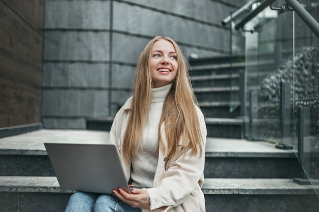 Feliz estudiante caucásica sentada en las escaleras cerca de la universidad con una computadora portátil, sonriendo y mirando hacia un lado. Mujer joven que trabaja en línea cerca de la oficina.