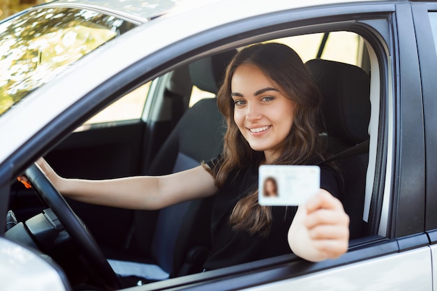 Foto feliz estudiante conductor sentado en el moderno coche plateado y mostrando el permiso de conducir