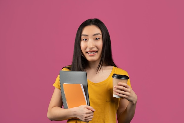 Feliz estudiante coreana sosteniendo cuadernos y café para llevar de pie sobre fondo violeta