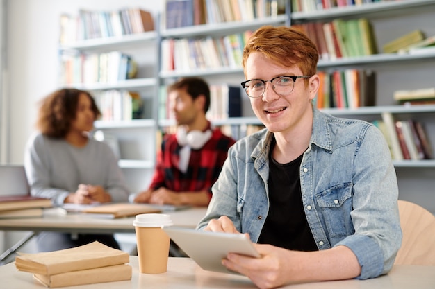 Feliz estudiante exitoso en ropa casual preparándose para el seminario mientras se desplaza en la tableta en el descanso