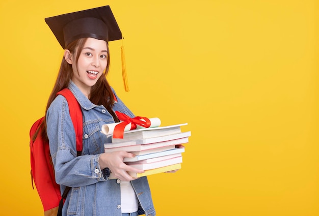 Feliz estudiante universitaria asiática con gorra de graduación sonriendo y sosteniendo libros