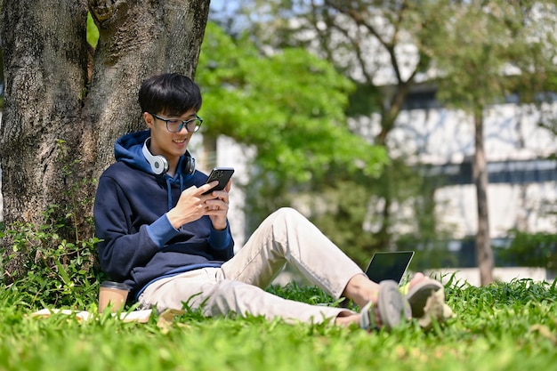 Feliz estudiante universitario asiático usando su teléfono inteligente debajo del árbol en el parque del campus