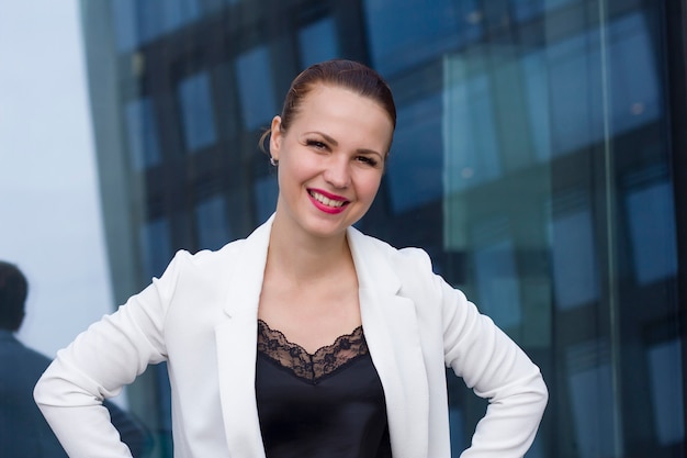 Feliz exitosa empresaria alegre en chaqueta está mirando a cámara y sonriendo con labios rojos. Trabajador de sexo femenino confidente con las manos en las caderas edificio de cristal de la oficina al aire libre. Vista frontal. Oficina exterior