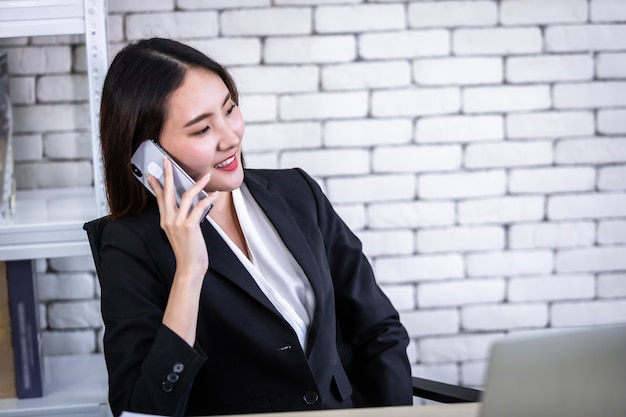 Feliz de exitosa joven empresaria asiática mantenga pick up smartphone trabajando con computadora portátil en mesa de madera blanca en fondo de oficina de pared blanca