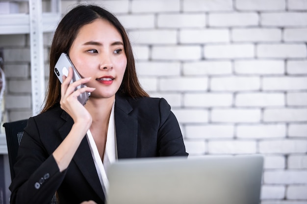 Feliz de exitosa joven empresaria asiática mantenga pick up smartphone trabajando con computadora portátil en mesa de madera blanca en fondo de oficina de pared blanca