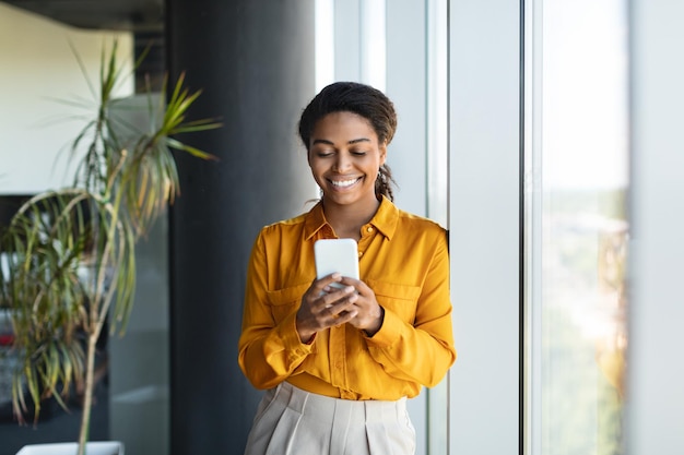 Feliz y exitosa mujer de negocios negra escribiendo en un teléfono inteligente de pie cerca de la ventana en el interior de la oficina moderna