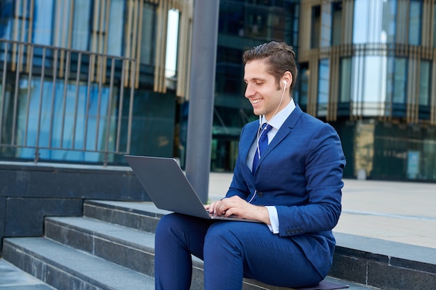 Feliz exitoso joven empresario trabajando en un portátil moderno, usando auriculares y sentado en las escaleras fuera de los rascacielos