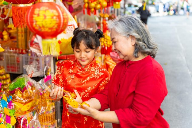 Feliz familia asiática abuela y nieta chica en ropa roja china eligiendo y comprando adornos decorativos para el hogar para la celebración del festival de vacaciones del Año Nuevo Lunar chino en el mercado de Chinatown