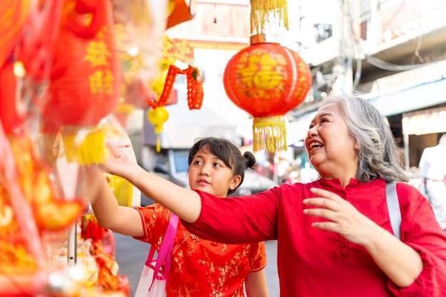 Feliz familia asiática abuela y nieta niña en vestido rojo eligiendo y comprando adornos decorativos para el hogar para la celebración del festival del Año Nuevo Lunar chino juntos en el mercado callejero de Chinatown