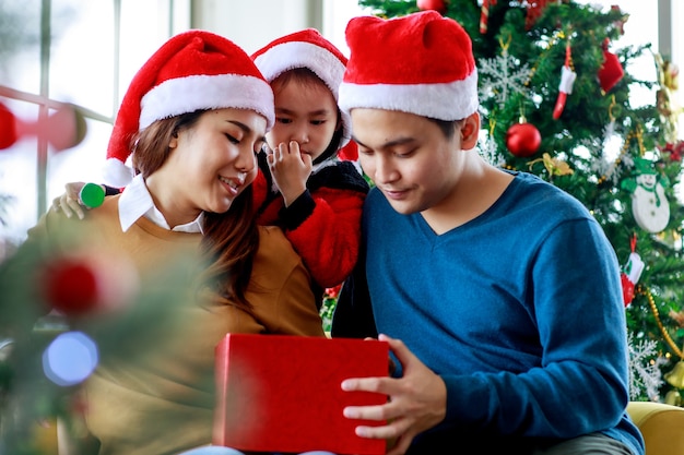 Feliz família asiática pai, mãe e filha usa uma camisola com chapéu de Papai Noel vermelho e branco, sorrindo, sorrindo, segurando a caixa de presente presente, prepare-se para desempacotar e abri-la juntos celebrando a véspera de Natal.