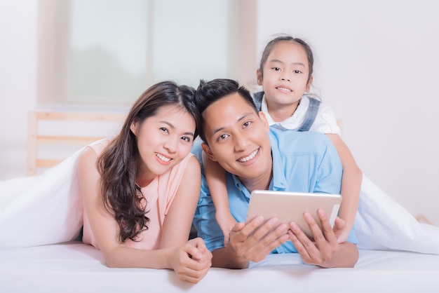 Feliz familia asiática relajante en la cama bajo una manta con tableta