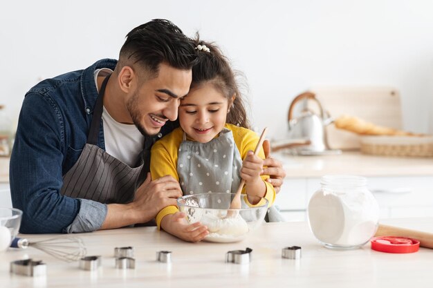 Feliz família do Oriente Médio pai e filha assando juntos na cozinha, menina árabe bonitinha misturando ingredientes para massa na tigela, gostando de cozinhar em casa com papai, closeup com espaço livre