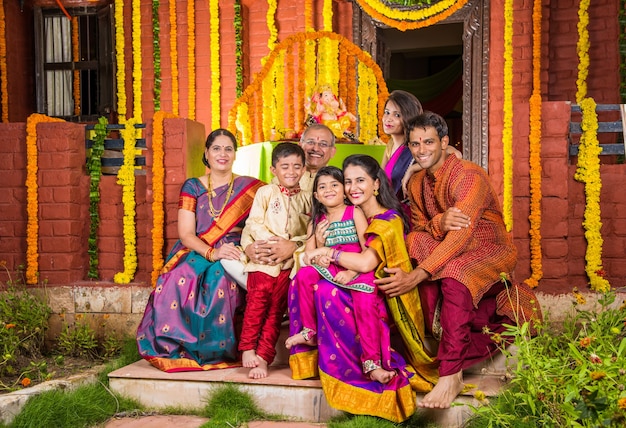 Feliz familia india celebrando el festival de Ganesh o Chaturthi dando la bienvenida o realizando Pooja y comiendo dulces con ropa tradicional en casa decorada con flores de caléndula
